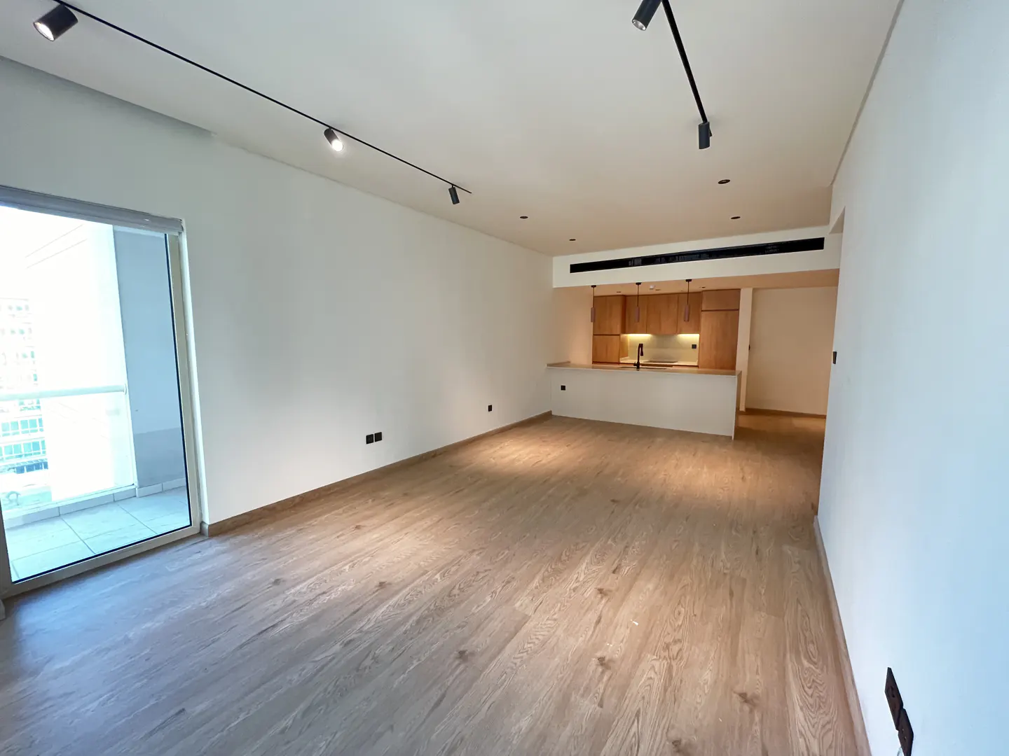 Modern open-plan living area with light wood floors, white walls, track lighting, and a kitchen with wood cabinets.