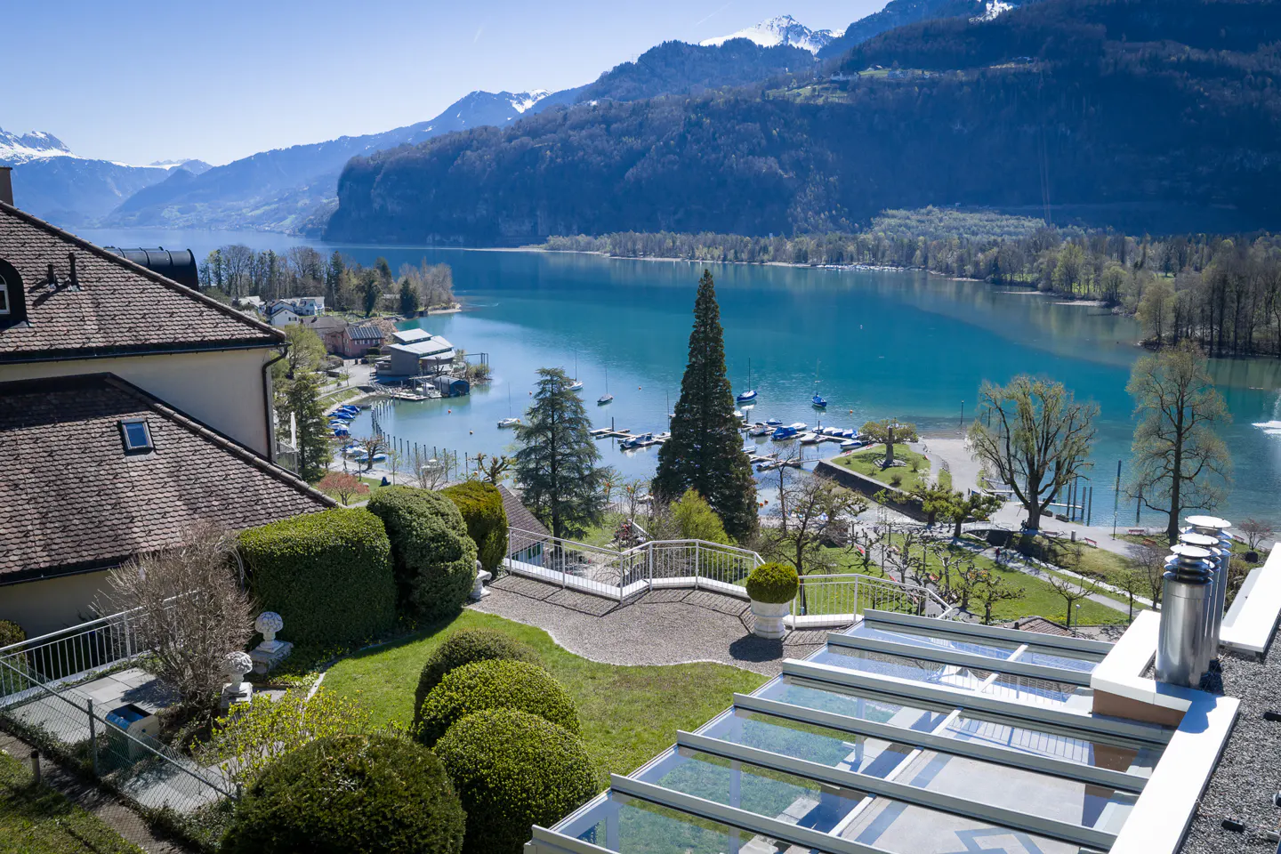 Scenic view from a house overlooking a turquoise lake, mountains, and a marina with boats. Green lawn and trees in the foreground.