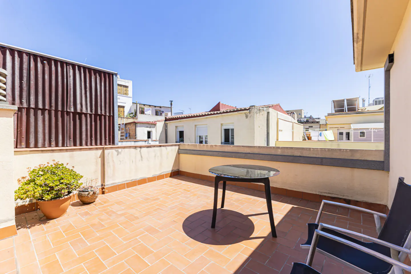 Outdoor patio with terracotta tiles, a round black table, chairs, and potted plants. Buildings are visible in the background under a clear blue sky.