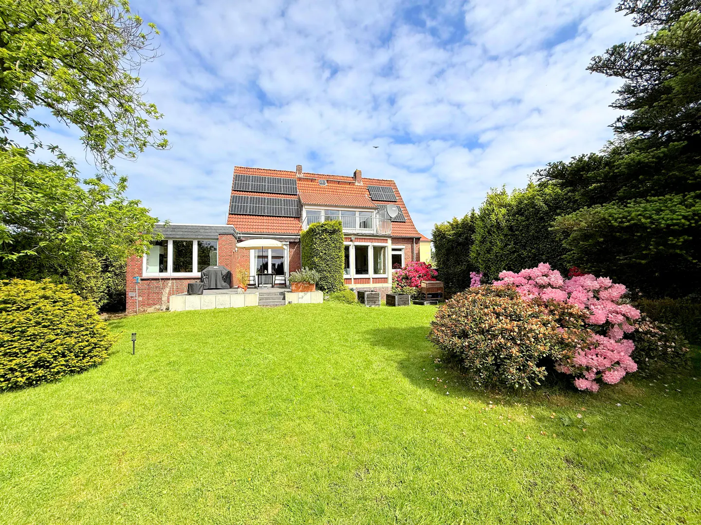 A red brick house with solar panels, a green lawn, and pink flowers under a blue, cloudy sky.
