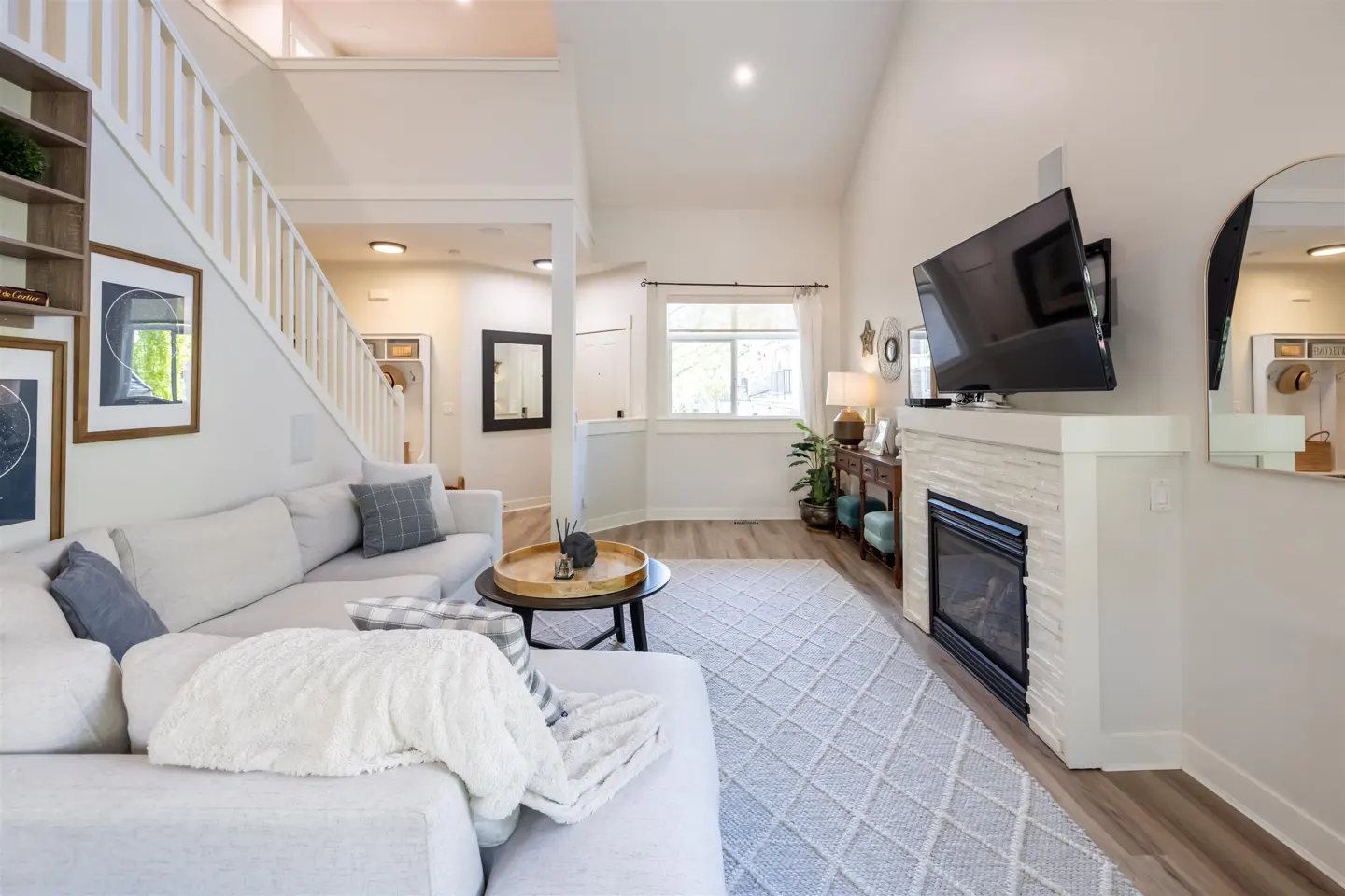 Bright living room with white sofa, fireplace, and TV. A staircase is visible on the left. The room has a light gray rug.