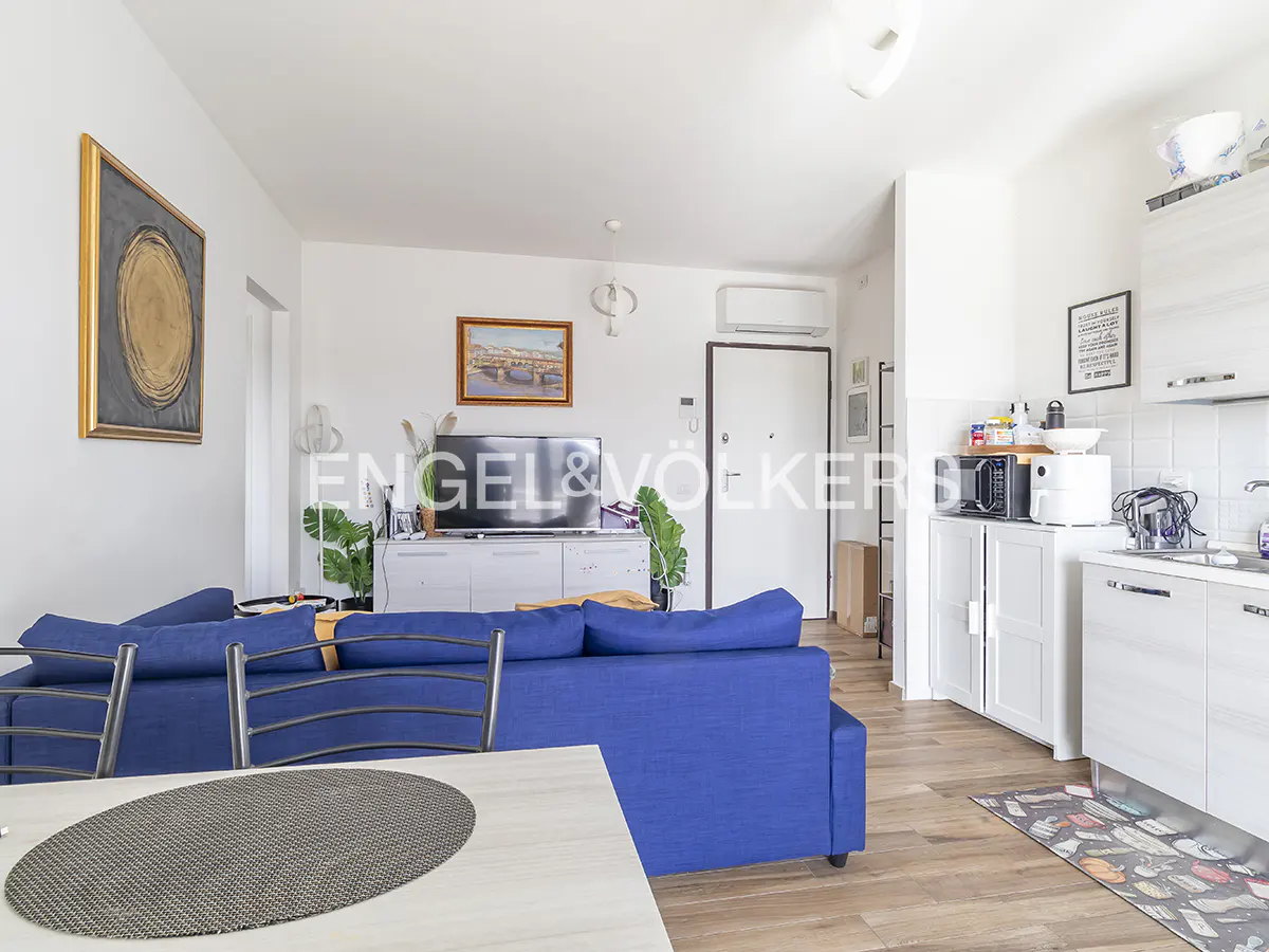 Bright apartment interior with blue sofa, white kitchen cabinets, and wood floors. Artwork adorns the walls.
