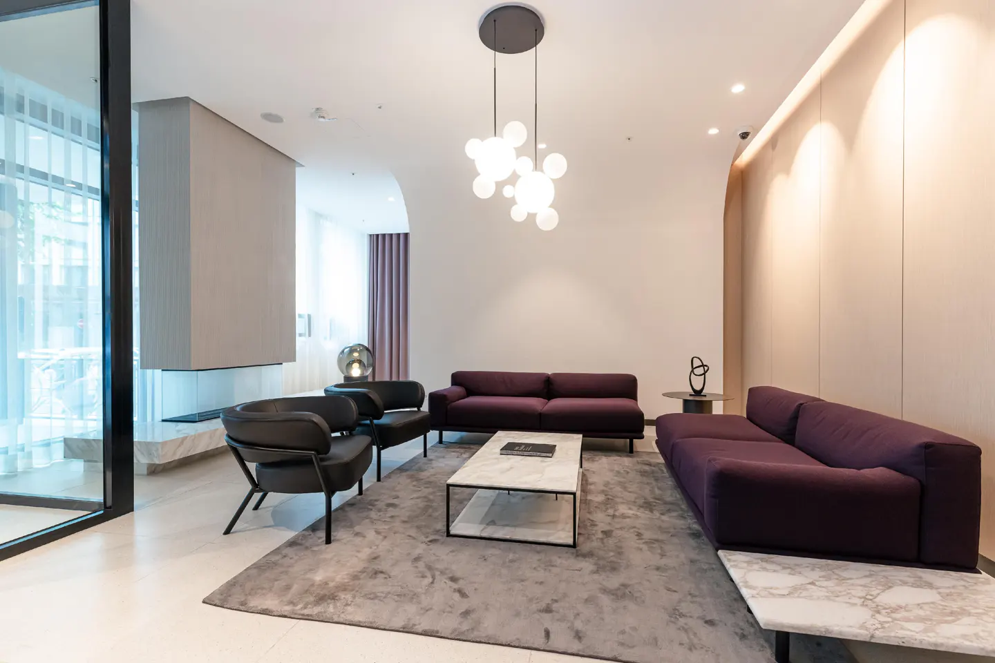 A modern living room with purple sofas, black chairs, and a marble coffee table on a gray rug. A unique light fixture hangs from the ceiling.