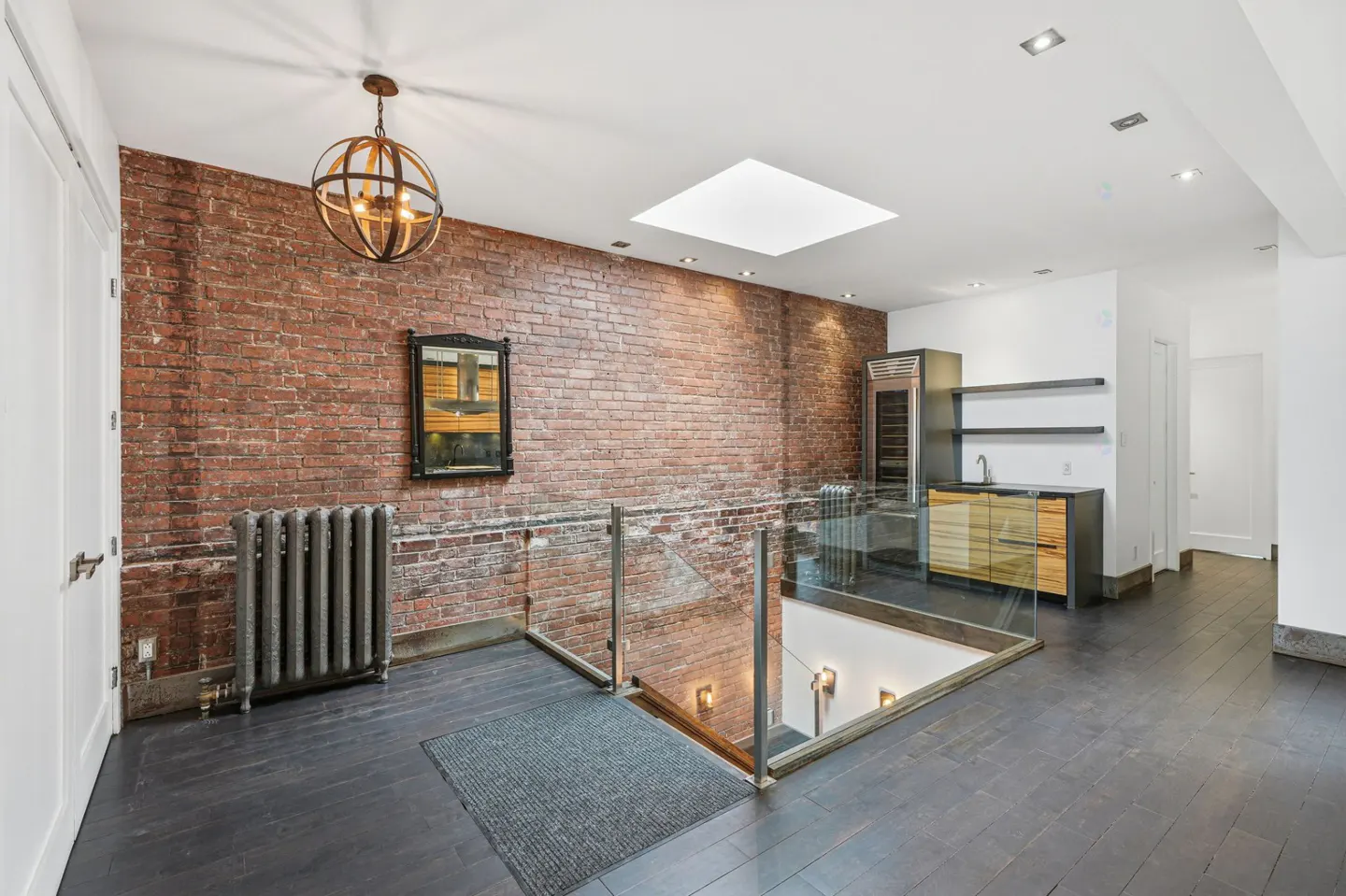Interior view of a loft with exposed brick, a skylight, and a glass railing overlooking a lower level. A modern light fixture hangs from the ceiling.