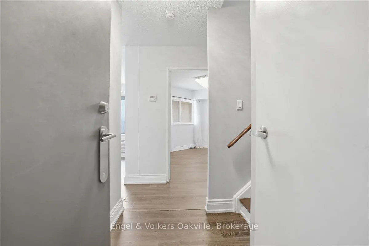 View through an open doorway into a bright, white hallway with light wood flooring and a staircase with a wooden handrail.