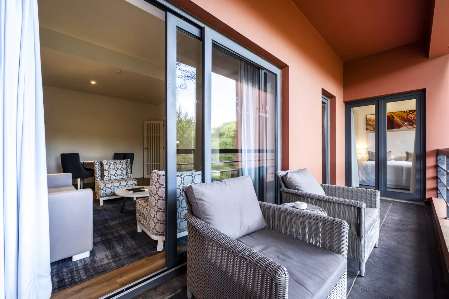 Balcony view of a hotel room with gray wicker chairs, cushions, and a small table. Sliding glass doors open to a living room with patterned chairs and a bedroom beyond.