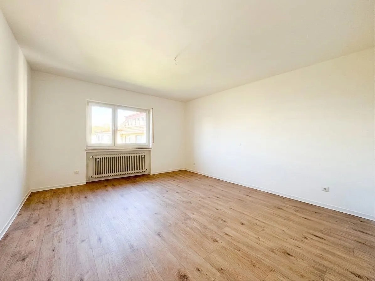 Bright, empty room with light wood floors, white walls, a window with a radiator, and a glimpse of a building outside.