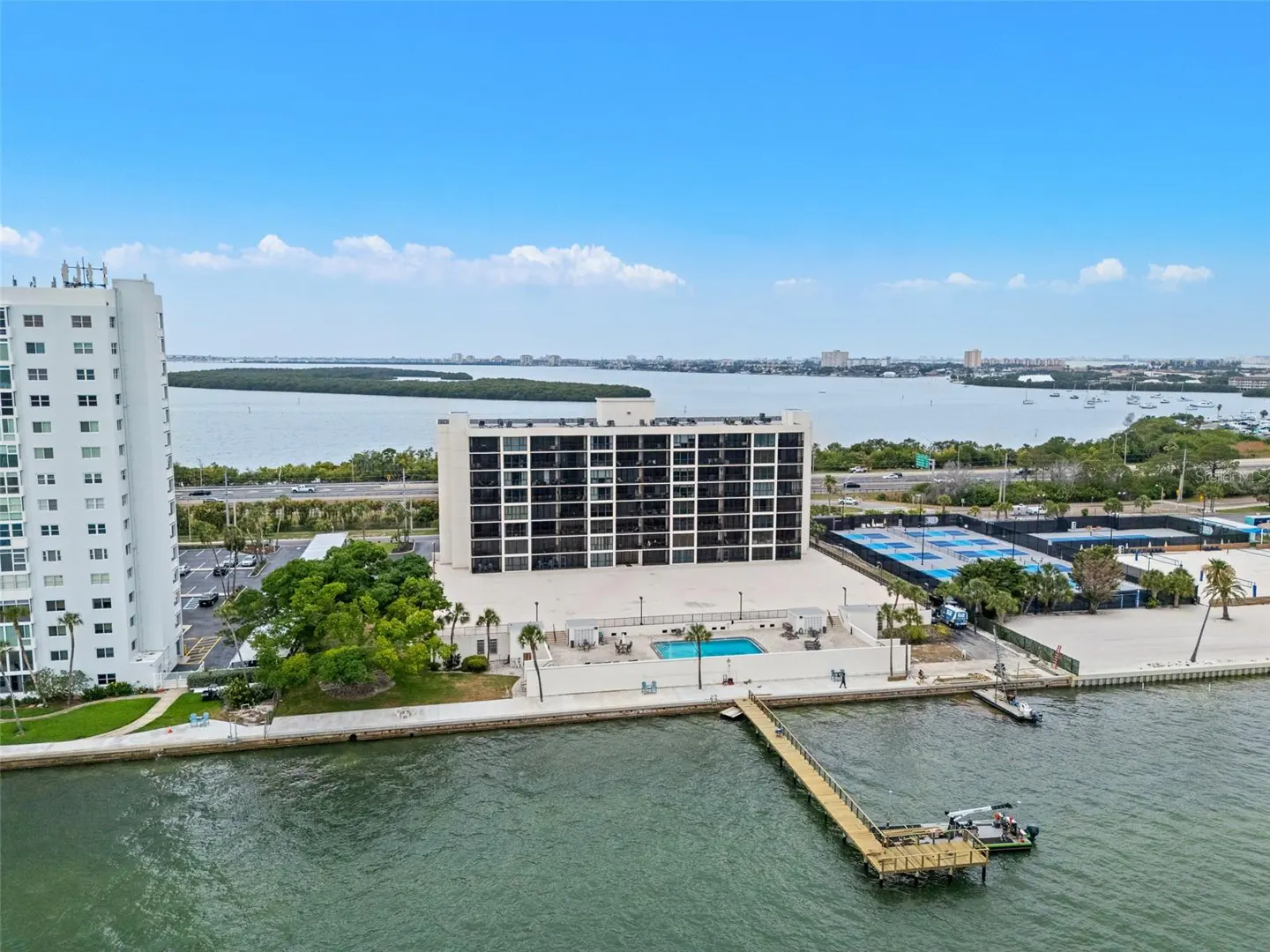 Aerial view of a waterfront condo with a pool, tennis courts, and a wooden dock extending into the water. Blue sky and bay in the background.