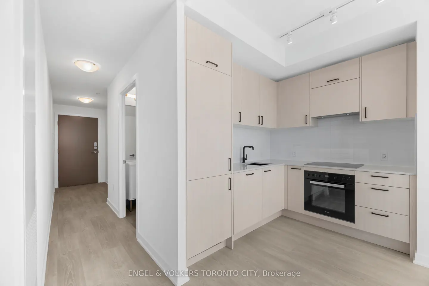 Modern kitchen with light beige cabinets, black hardware, and stainless steel appliances, adjacent to a hallway.