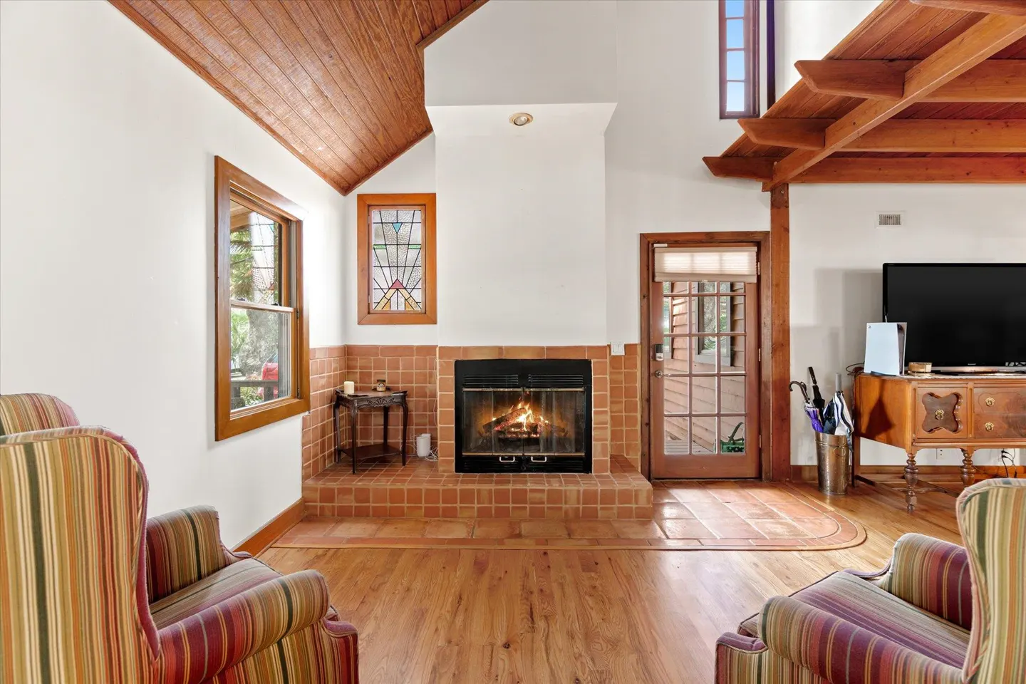 Living room with a fireplace, wood floors, and striped armchairs. A stained glass window is above the fireplace. A TV sits on a wooden dresser.