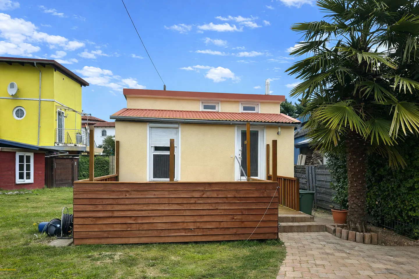 A tan house with a brown deck and a red roof, next to a palm tree under a blue, cloudy sky.