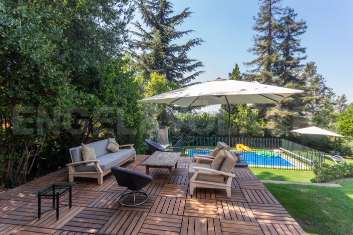 Outdoor patio with wood tile flooring, sofa, chairs, and coffee table. A pool is visible in the background, surrounded by green grass and trees.