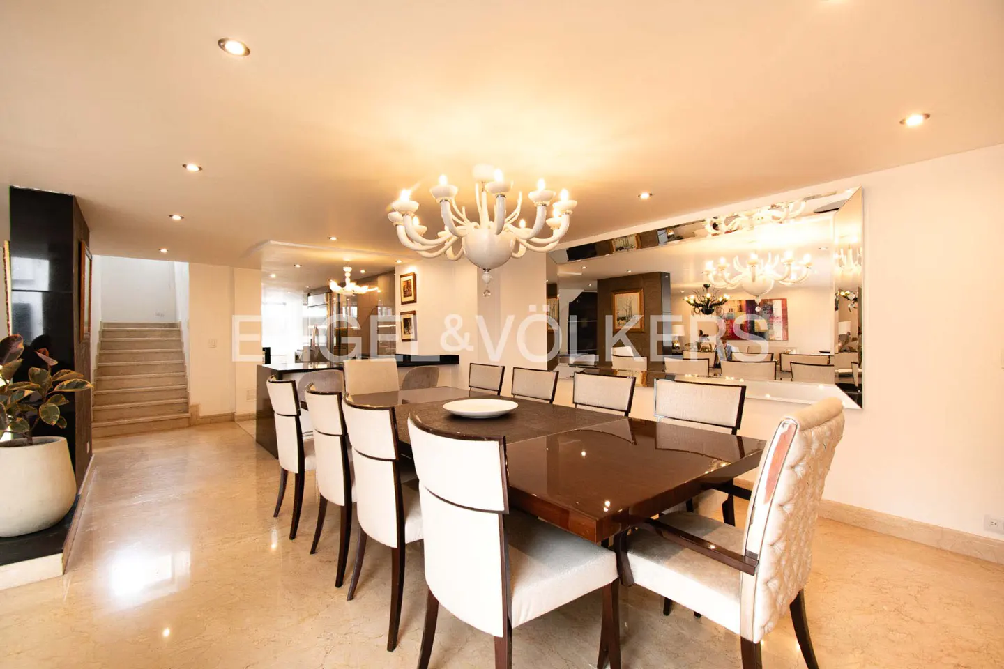 Bright dining room with a long, dark wood table, white chairs, and a white chandelier. A large mirror reflects the room. Marble floors and stairs are visible.