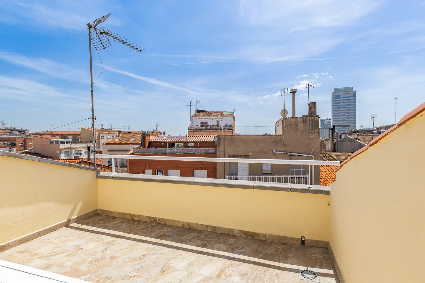 A rooftop terrace with beige walls and tiled floor overlooks a cityscape under a blue sky. An antenna stands tall against the skyline.