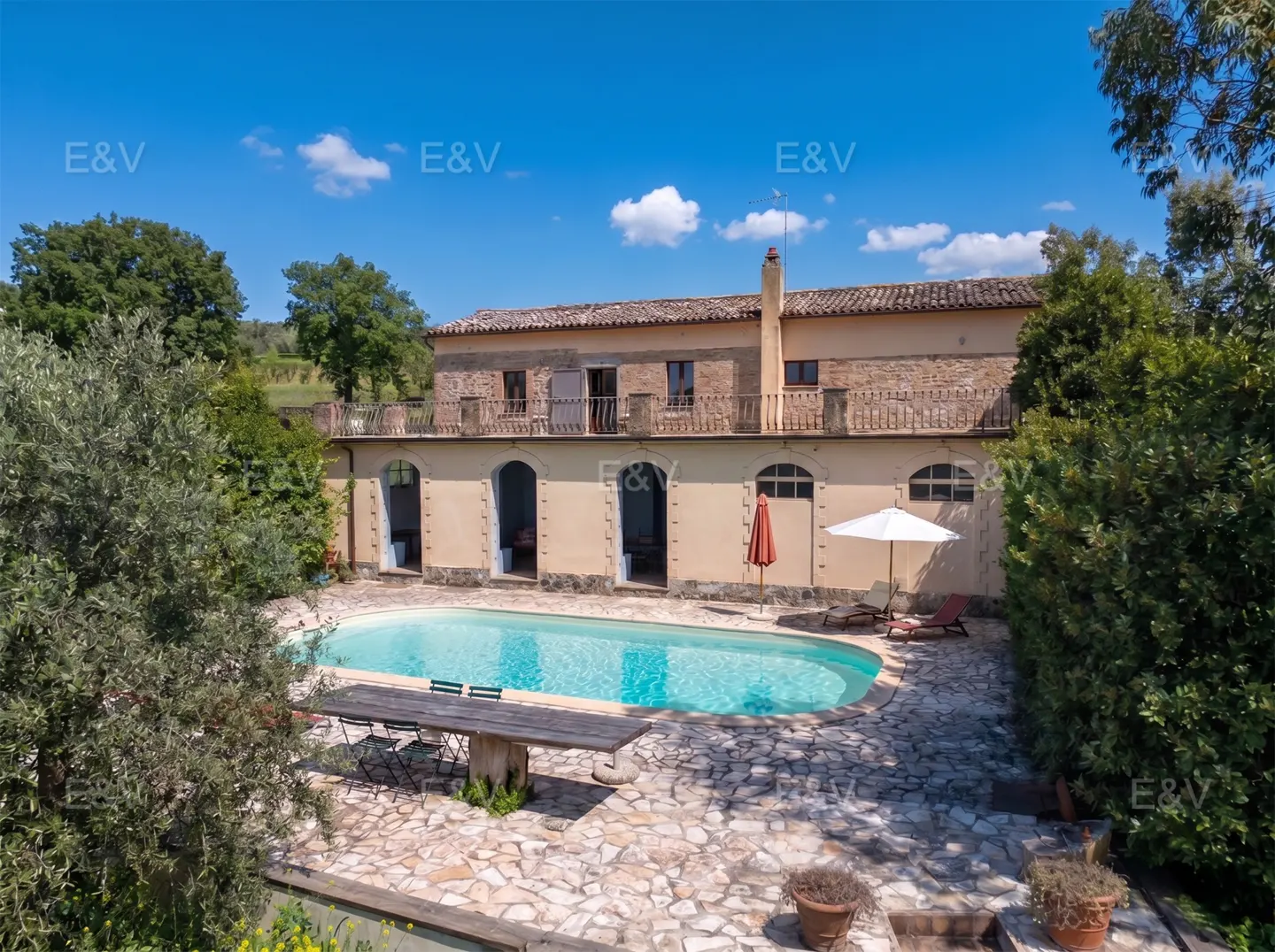 Exterior view of a two-story beige house with a pool, stone patio, table, chairs, and umbrellas on a sunny day.
