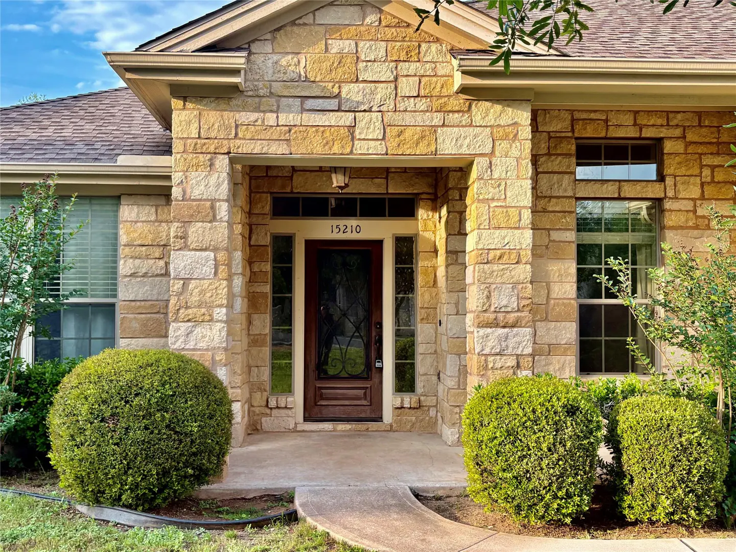 Front exterior of a tan stone house with a brown front door and manicured green bushes.