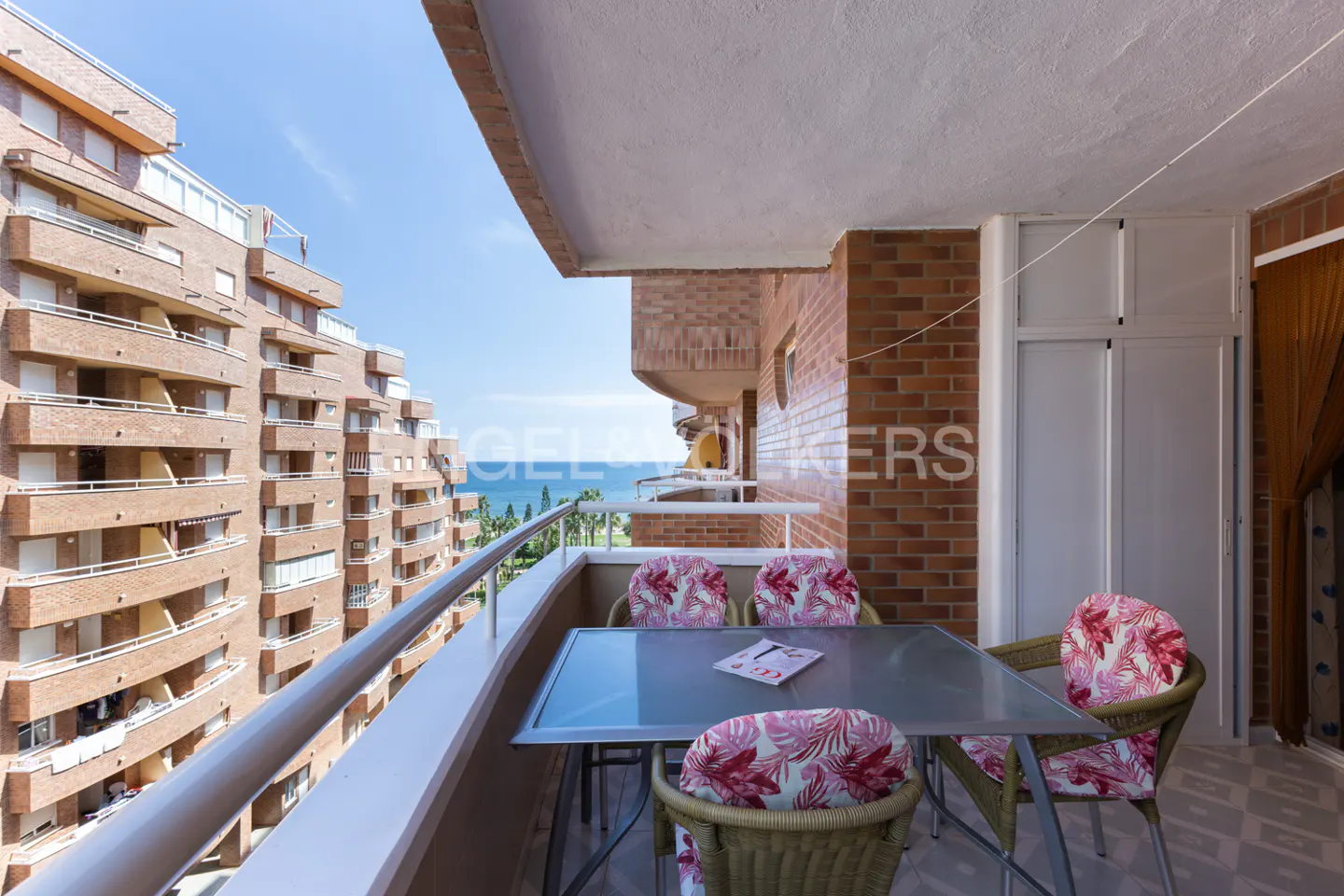 Balcony view with table, chairs, and ocean. Brick building and blue sky in the background.