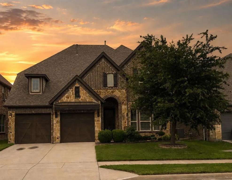 Two-story brick and stone house with a brown roof, two-car garage, and green lawn at sunset.