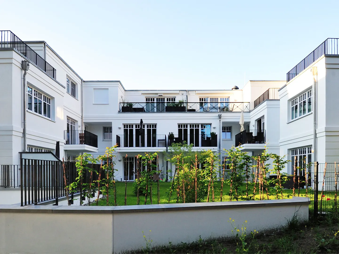 Exterior view of a modern, white apartment building with black balconies and a green courtyard.