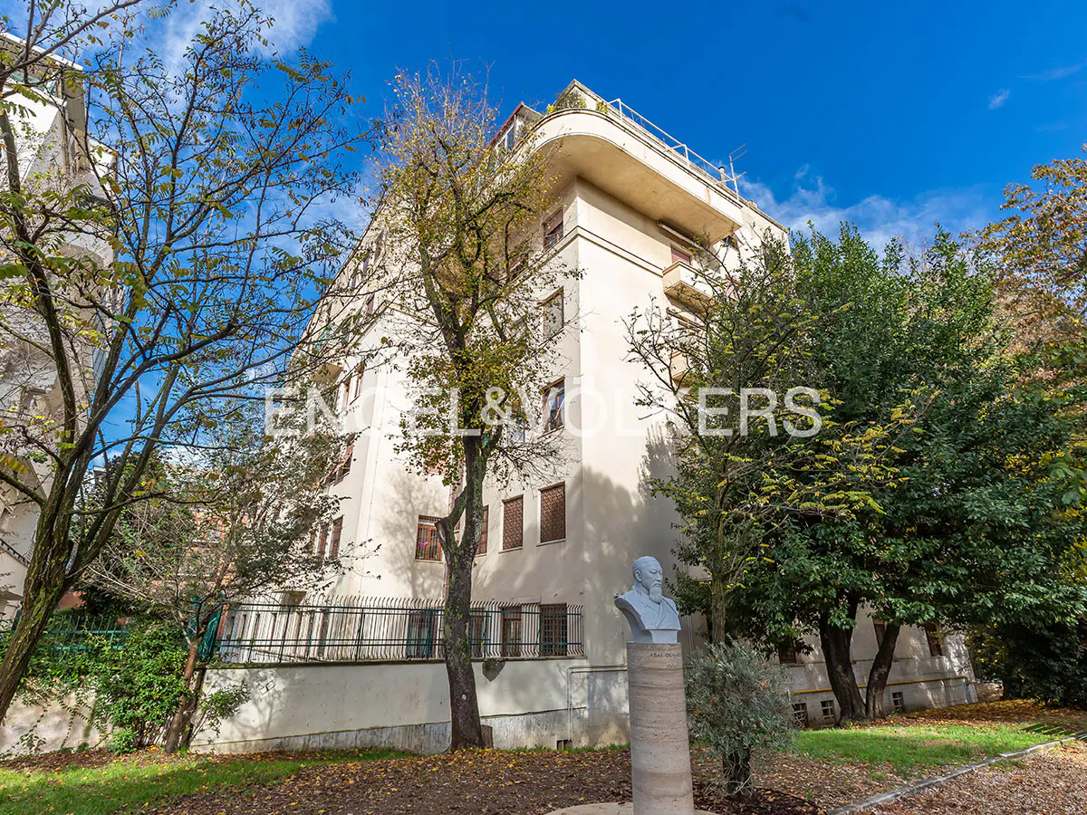 Exterior view of a multi-story white building with trees, a bust statue, and a blue sky.