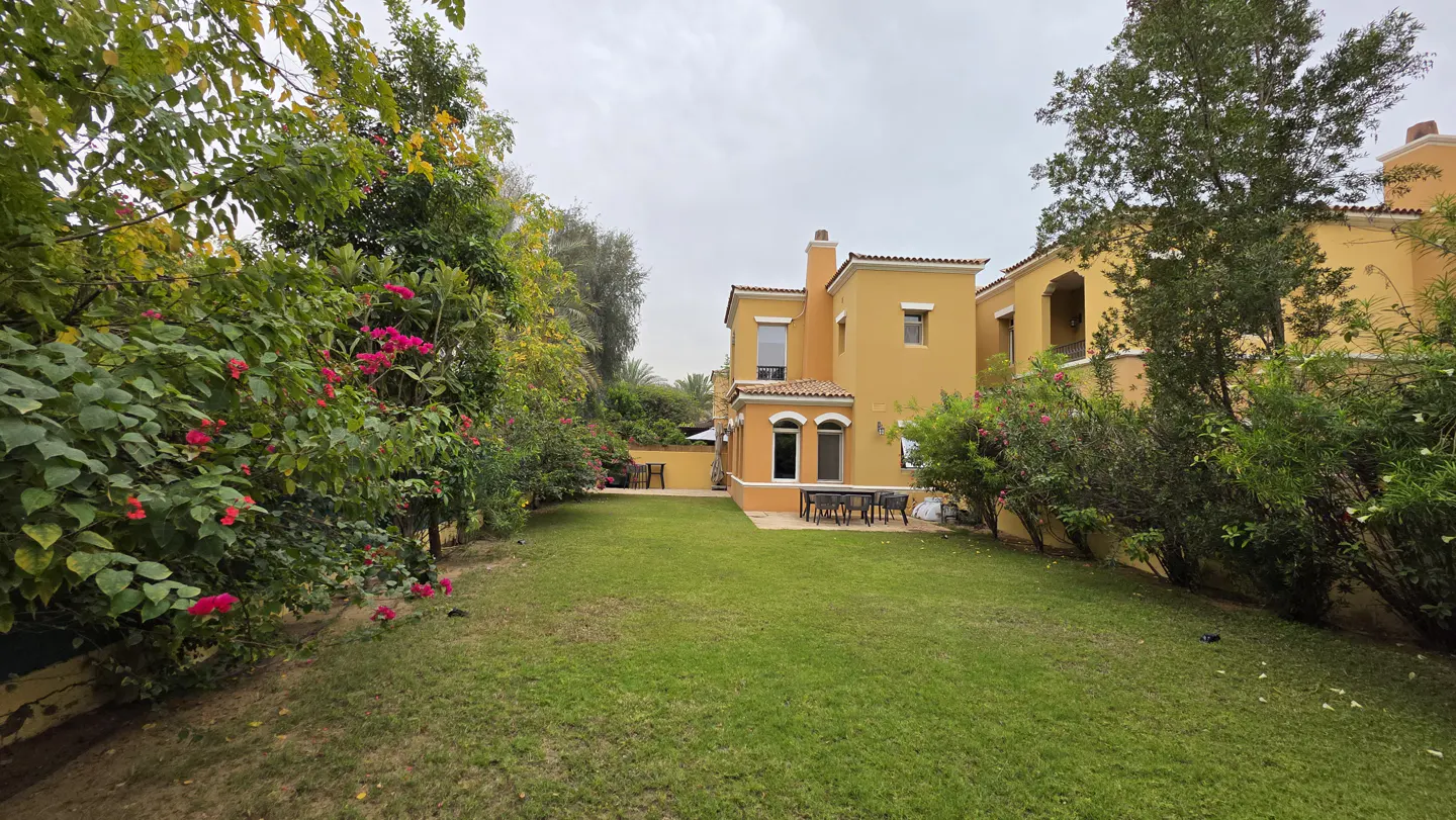 A lush green backyard with a patio set, surrounded by flowering bushes and trees, leading to a yellow two-story villa.