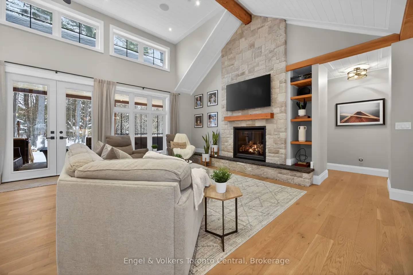 Living room with stone fireplace, TV, and built-in shelves. Large windows overlook a snowy landscape. Beige sofa and wood floors.