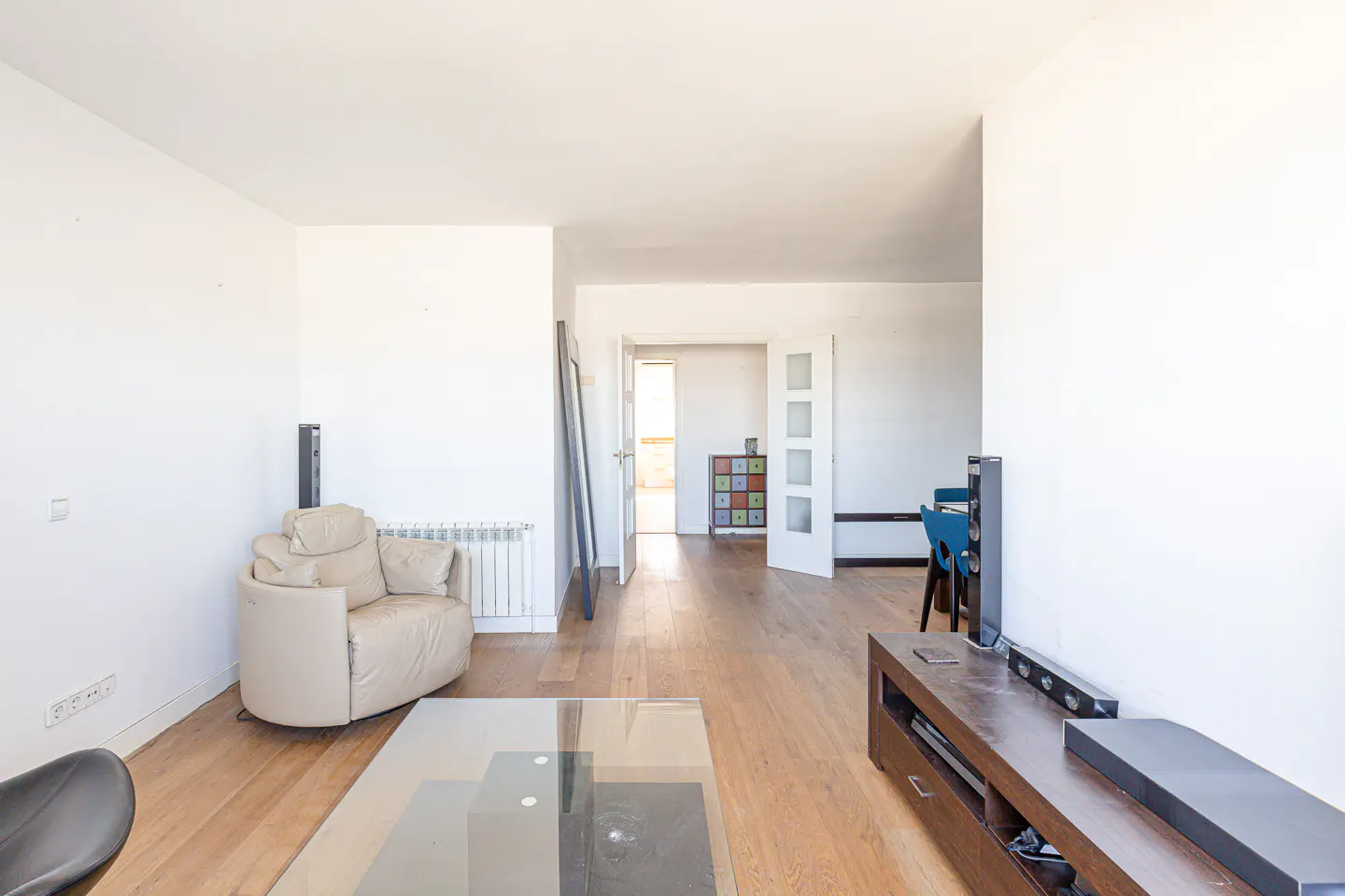 Bright living room with hardwood floors, white walls, and a beige armchair. A glass coffee table sits in front of a wooden TV stand. Open doorway leads to another room.