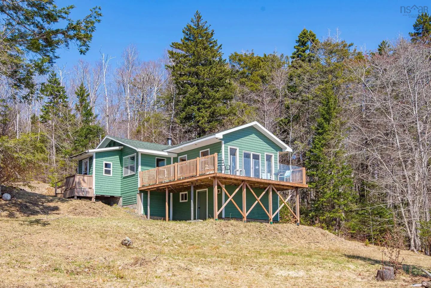 Green house with a wooden deck on a grassy hill, surrounded by trees under a blue sky.