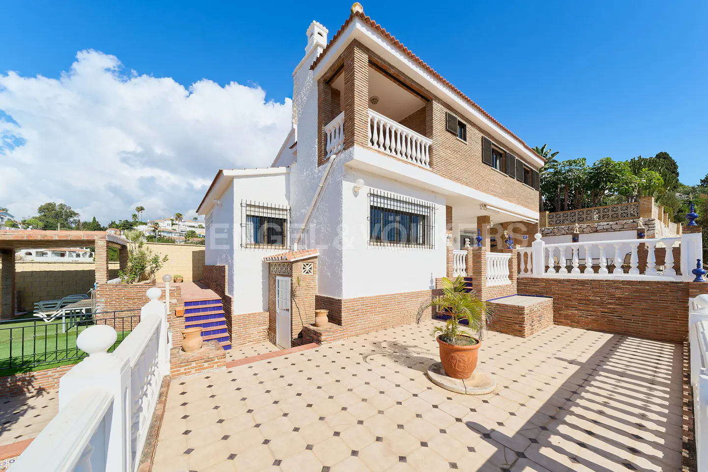 Exterior view of a two-story house with white walls, brick accents, and a tiled patio under a blue sky.