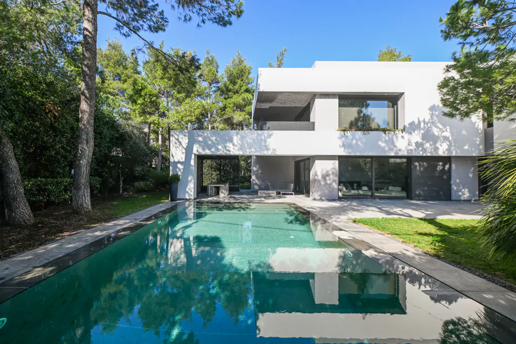 Modern white house with a turquoise pool reflecting trees and sky. Large windows and an open-air patio are visible.