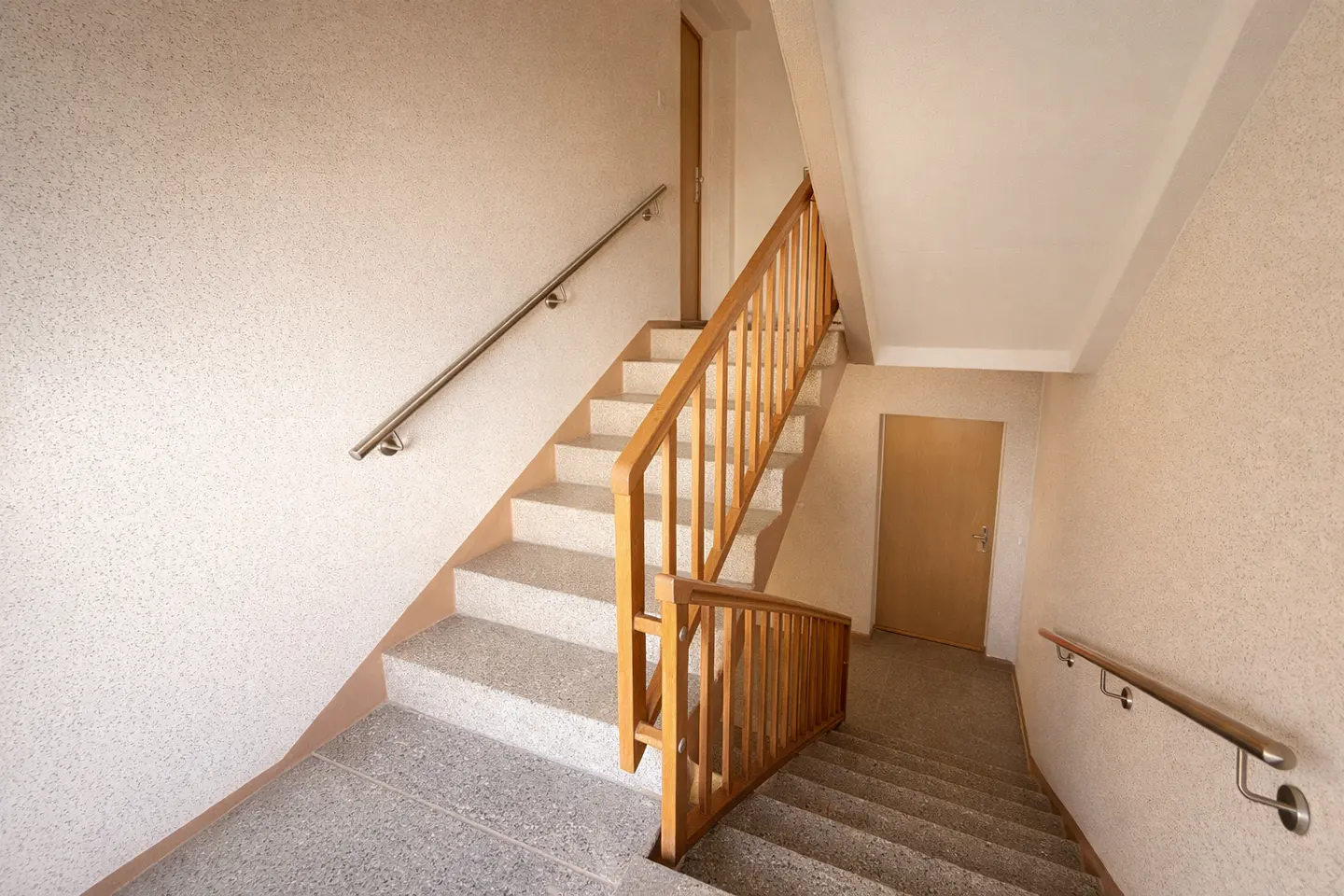 Beige stairwell with speckled walls, featuring a wooden railing and stainless steel handrail. A closed door is visible on the landing.