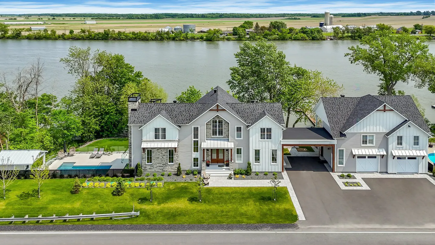 Aerial view of a modern gray and white farmhouse-style home with a pool, near a river and farmland.