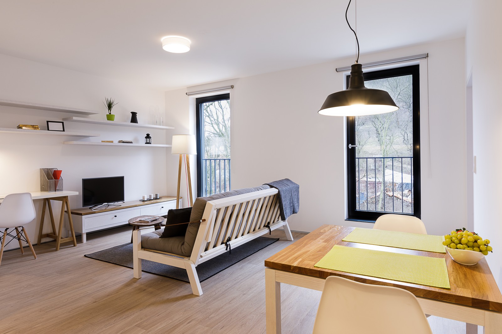 Modern open-plan living room with a sofa, dining table, desk, and large windows overlooking a wooded area.