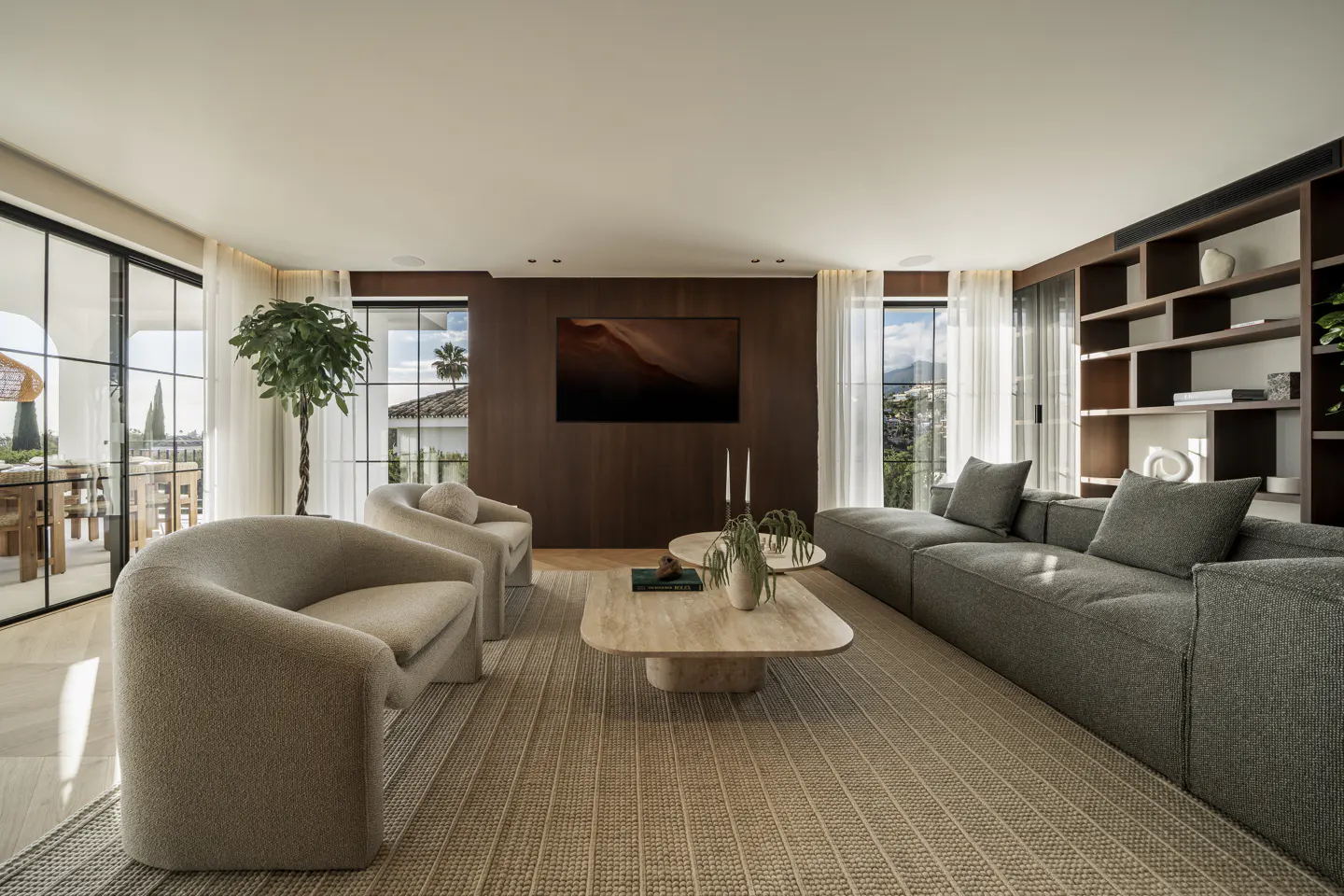 A modern living room with beige chairs, a gray sofa, and a light brown rug. A large TV hangs on a dark wood wall. Windows offer a view of the outdoors.