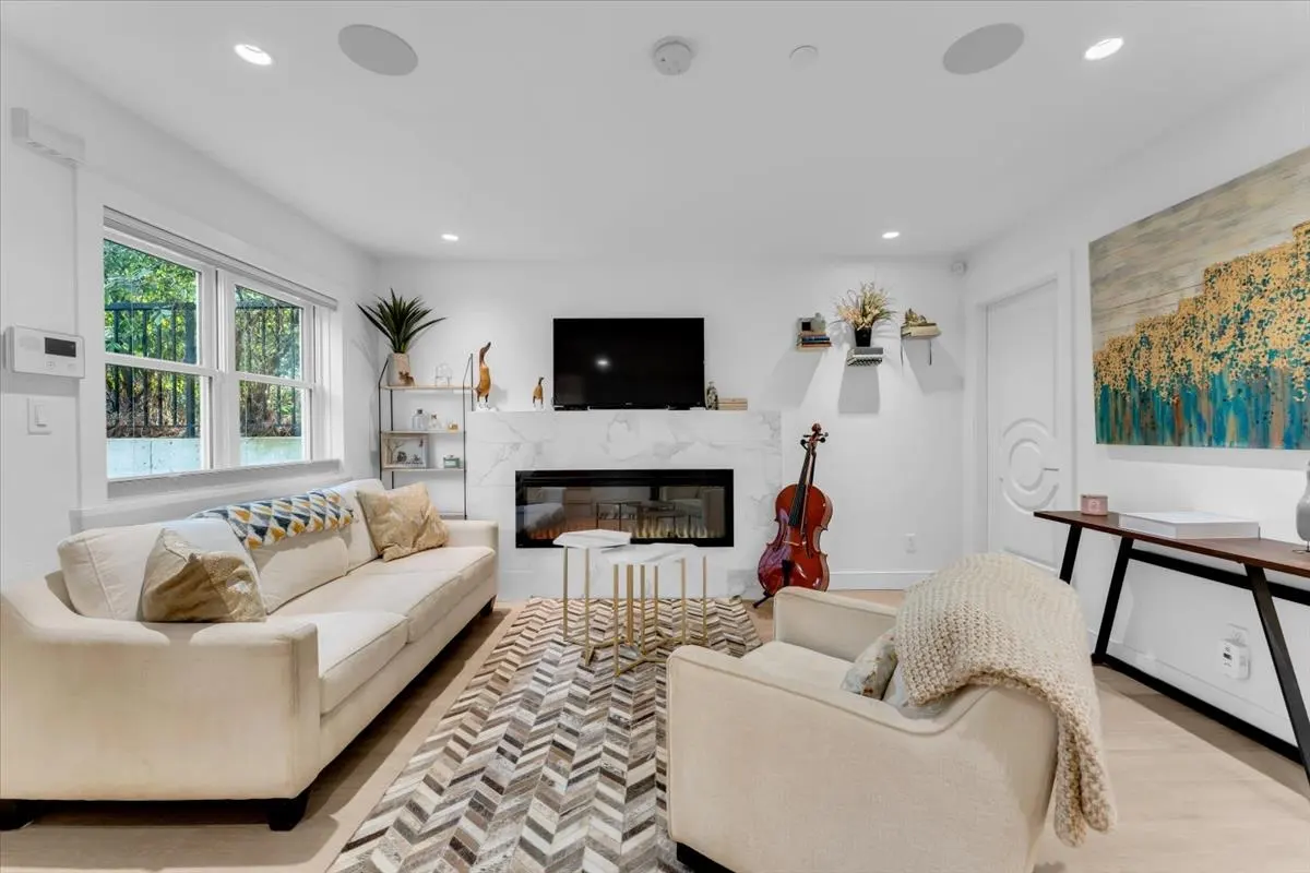 Bright living room with white walls, a fireplace, and a TV. Beige sofa and armchair face each other over a patterned rug. A cello stands near the fireplace.