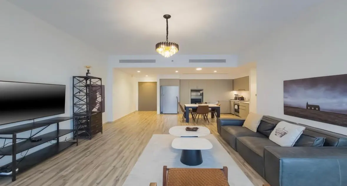 A modern apartment living room with a gray sofa, white coffee tables, and a TV. The kitchen is visible in the background.