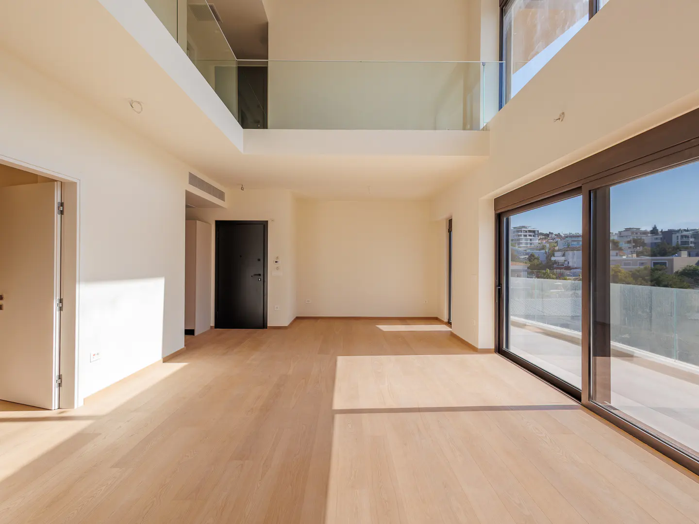 Bright, empty room with light wood floors, white walls, and a black door. Large sliding glass doors lead to a balcony with a city view.