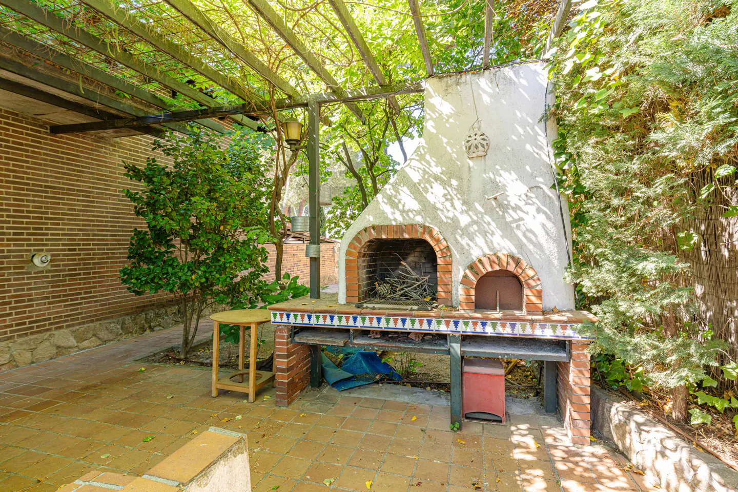 Outdoor patio with a white brick barbecue, tiled counter, wooden stool, and lush greenery under a wooden pergola.