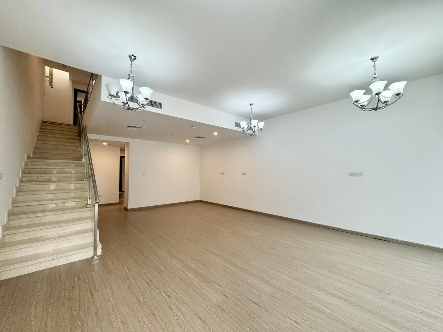 Empty living room with light wood floors, white walls, and three chandeliers. A staircase with metal railing is on the left.