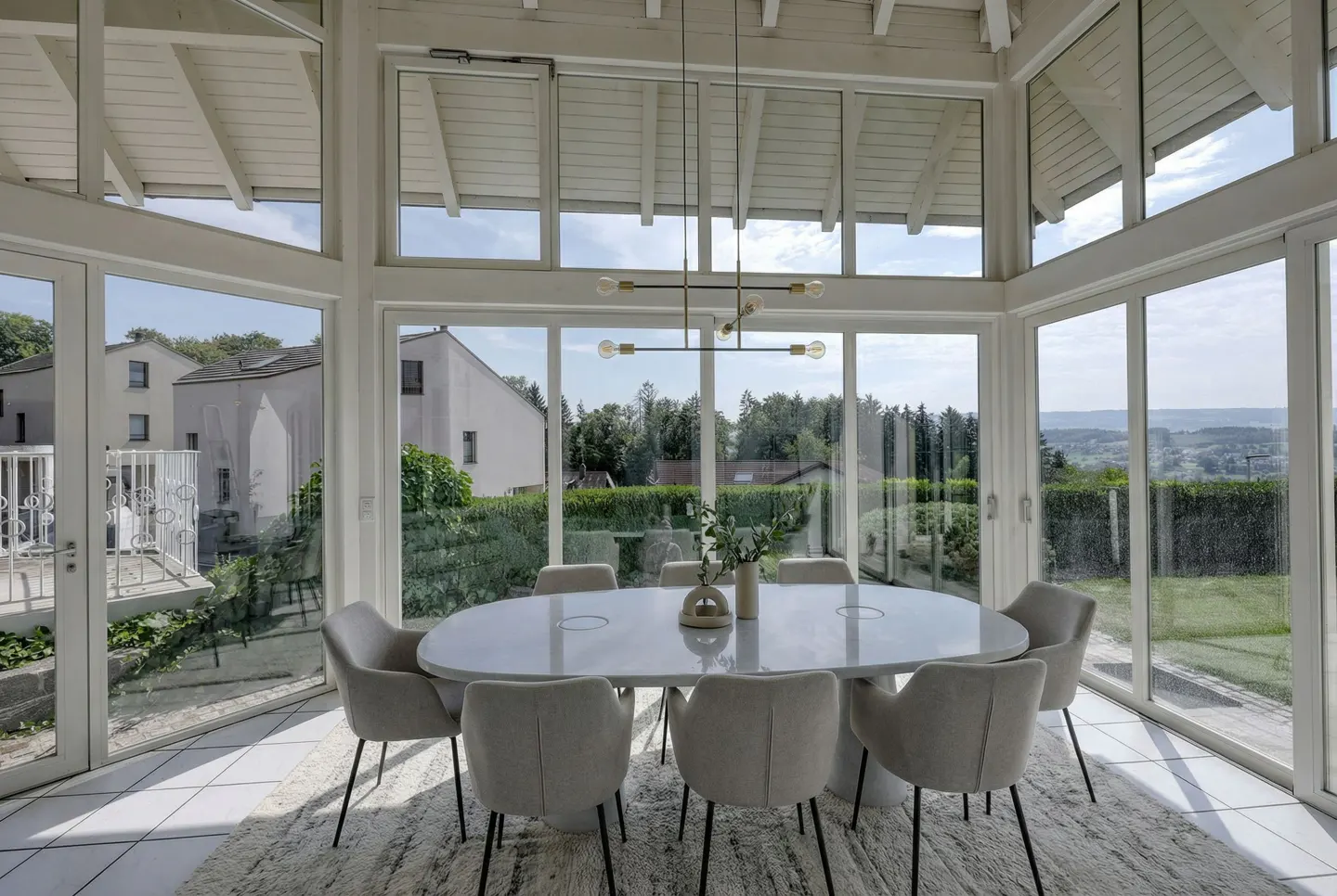Bright sunroom with white-framed windows, an oval table with eight chairs, and a modern chandelier. Outside, there are green trees and houses.
