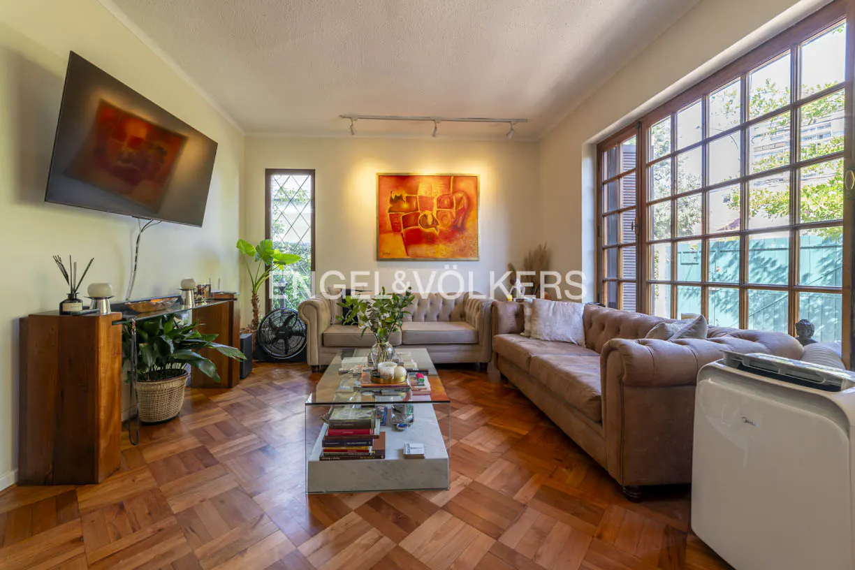 Living room with parquet floors, brown sofas, glass coffee table, and large windows. A TV hangs on the wall.