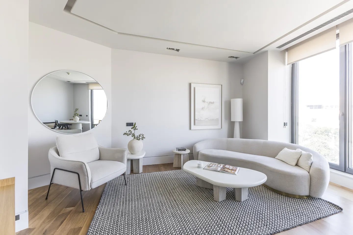 Bright living room with white walls, a curved sofa, chair, and a round mirror reflecting a dining area. A patterned rug covers the wooden floor.
