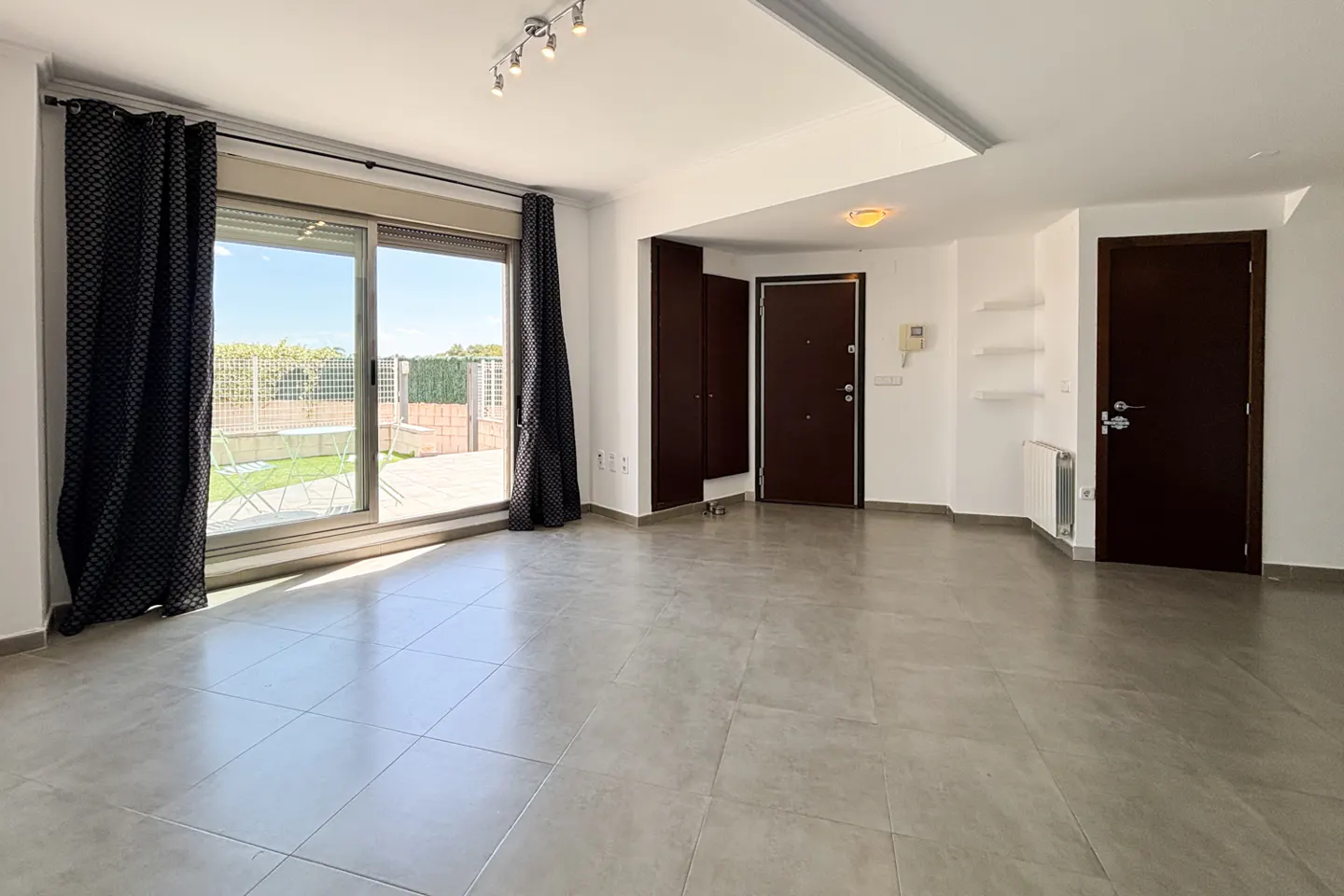Bright, empty room with gray tile floor, white walls, and dark brown doors. Sliding glass door to patio with table and chairs.