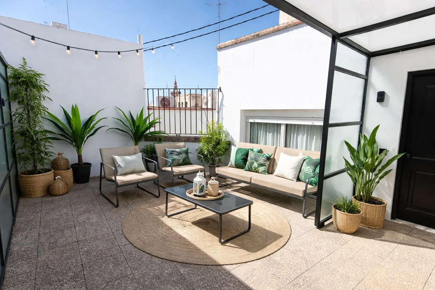 Outdoor patio with a beige sofa, two chairs, a black coffee table on a round jute rug, and potted plants against a white wall.