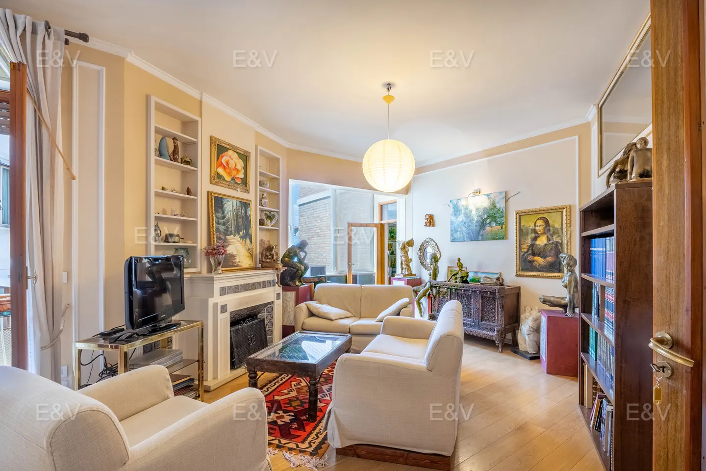 A bright living room with white sofas, a fireplace, and art. Bookshelves and a large, round light fixture add to the room's charm.