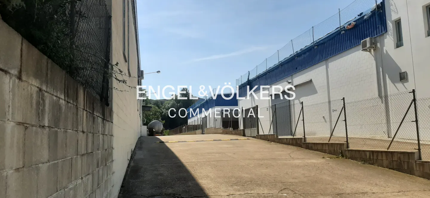 Concrete driveway between two industrial buildings with white walls and blue metal trim under a clear blue sky.