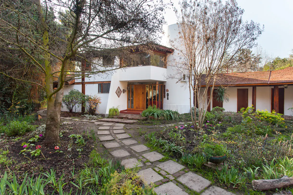 A white two-story house with a brown roof and a stone path leading to the front door. The house is surrounded by trees and plants.