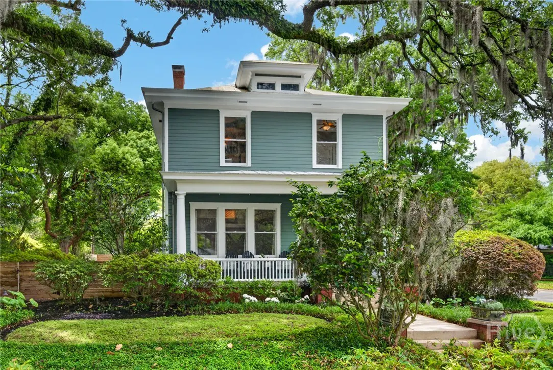 Two-story teal house with white trim, a front porch, and a green lawn surrounded by trees.