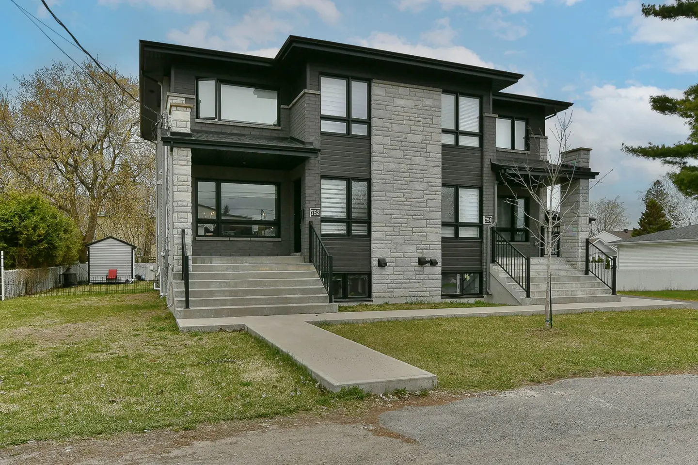 Modern two-story duplex with gray stone and dark siding, concrete steps, and black railings on a grassy lawn.