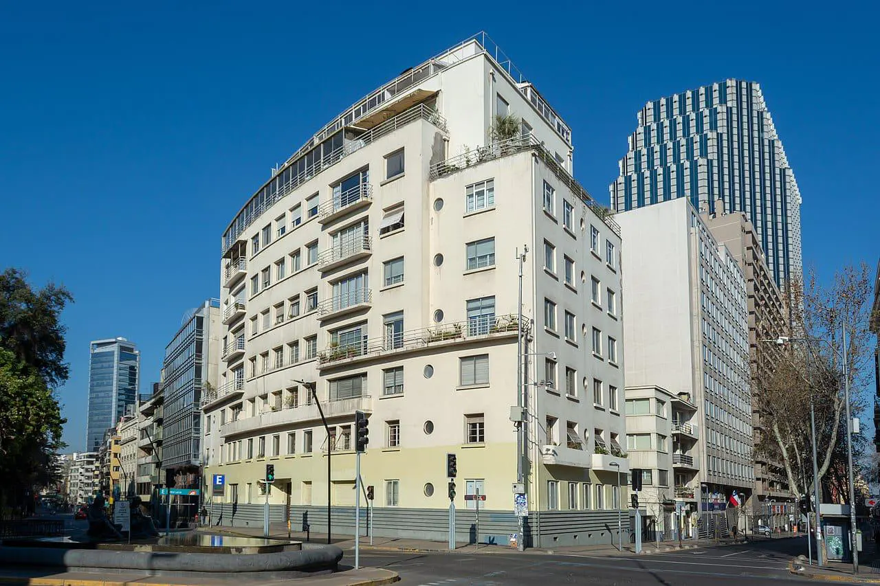 Street view of a multi-story, cream-colored apartment building with balconies, and a modern skyscraper in the background.